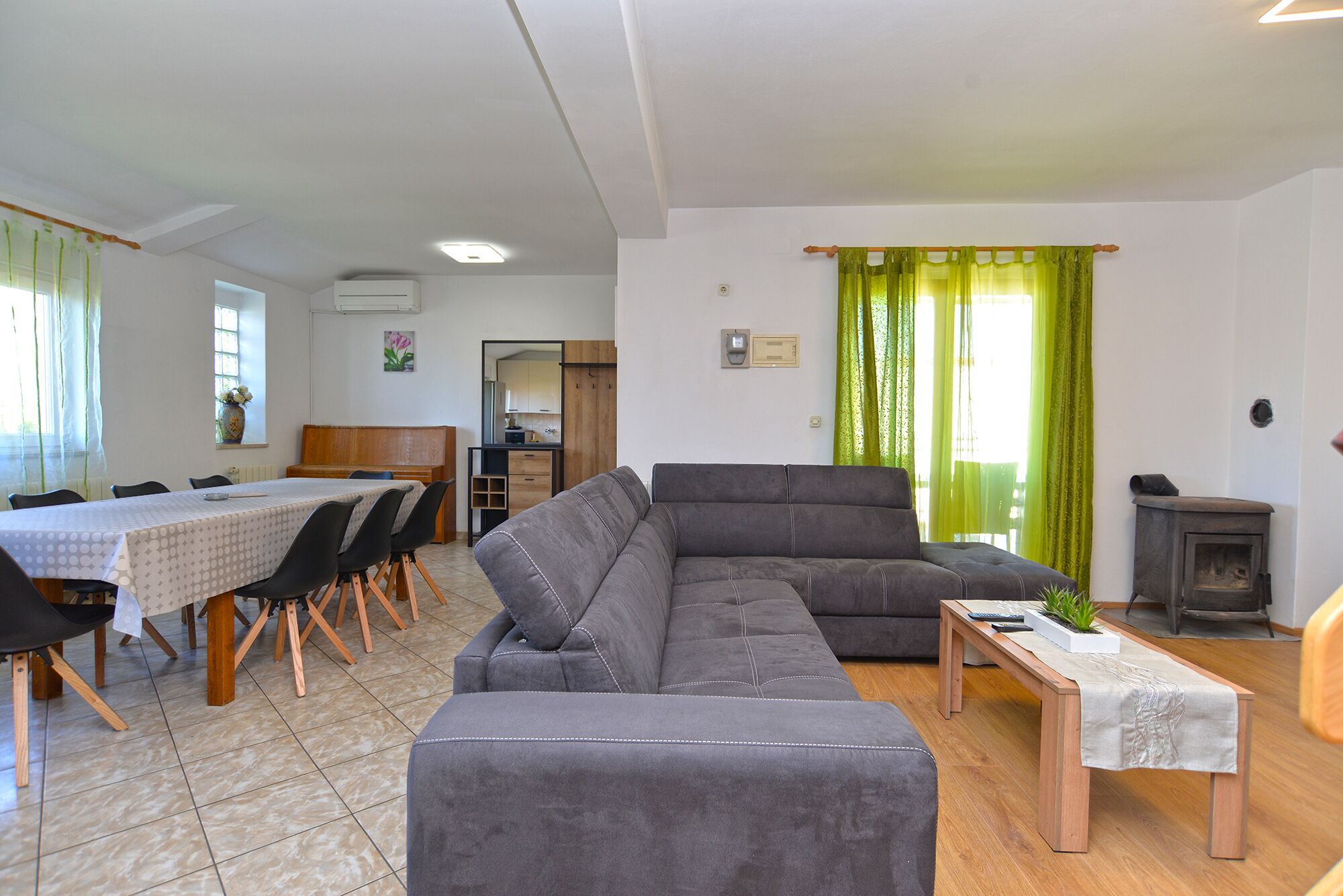 A living room with a grey sofa, a dining table with chairs, a small wooden table, and a green curtain.