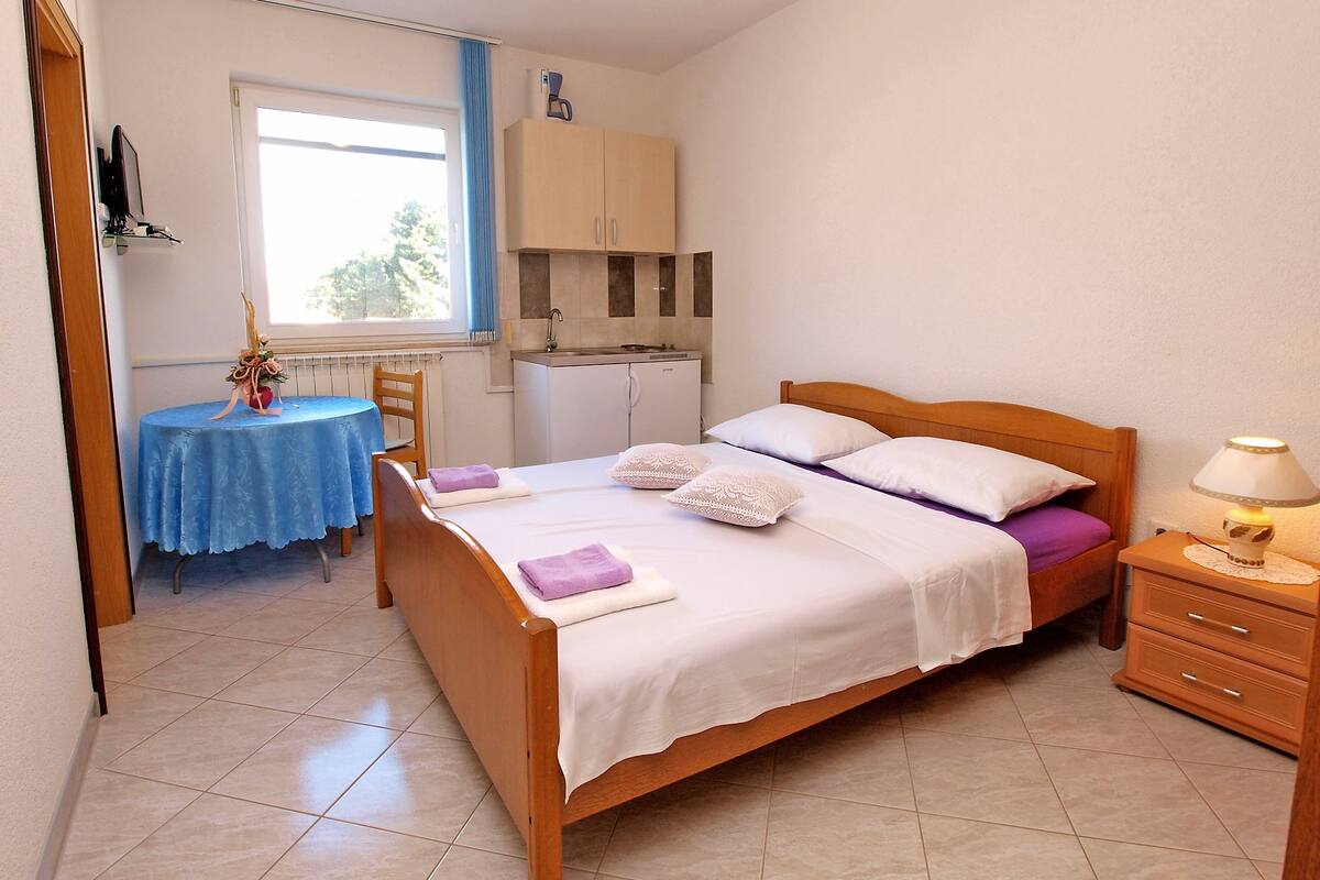 A bedroom with a wooden bed, a small table with a blue tablecloth, a kitchenette, and a window with curtains.