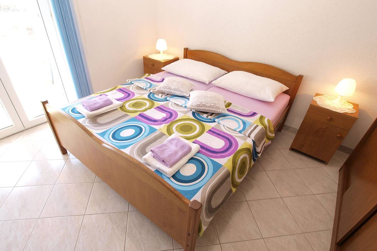 A bed with a colorful circular-patterned bedspread, two bedside tables with lamps, and a window with a blue curtain.