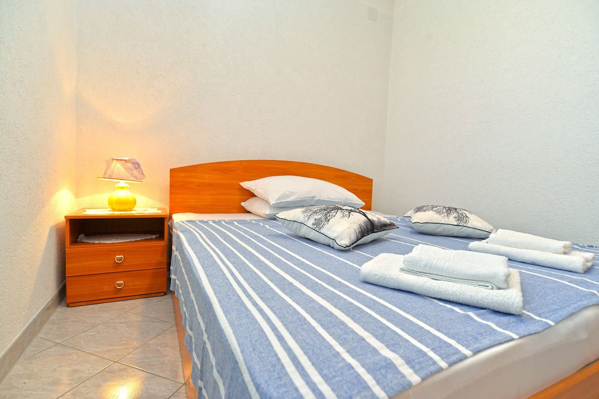A neatly made bed with striped bedding, a wooden headboard, and two pillows. There is a nightstand with a lamp and a drawer.