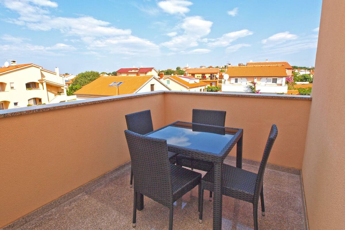 Un balcón con una mesa de vidrio y cuatro sillas de mimbre, con vistas a una zona residencial con casas de techos anaranjados.