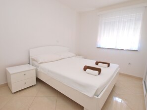 Ein sauberes, weißes Schlafzimmer mit einem Bett, einem Nachttisch und einem Fenster mit durchsichtigen Vorhängen.