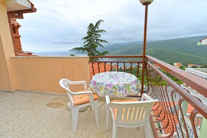 A balcony with a round table and two chairs, overlooking a coastal landscape with a view of the sea and hills.