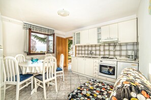 Une cuisine compacte avec des meubles blancs, une cuisinière à gaz et une machine à café. On y trouve un coin repas avec une table et des chaises, et une fenêtre offre une vue sur la mer.