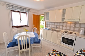 Une cuisine compacte avec des meubles blancs, une table à manger avec des chaises, une fenêtre avec des rideaux et une vue sur la verdure à l’extérieur.