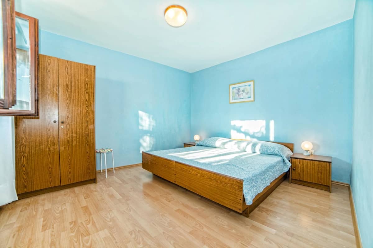 Een slaapkamer met een houten bed, nachtkastje, een klein krukje en een kledingkast.