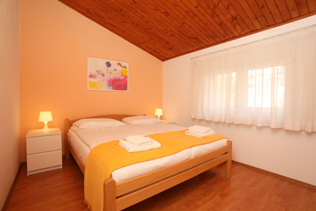 Een slaapkamer met een houten plafond, een bed met wit beddengoed, twee bedlampen, een raam met witte gordijnen en een kleurrijk abstract schilderij aan de muur.