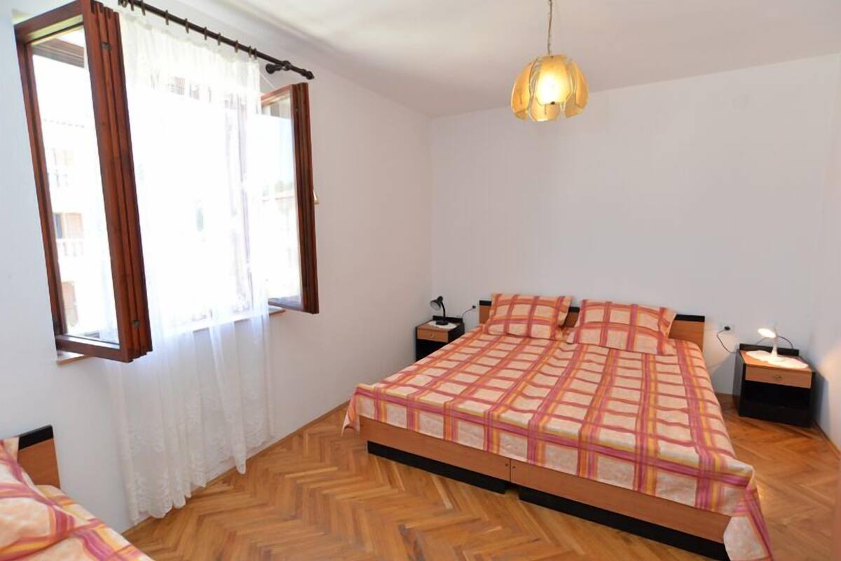 A bedroom with a wooden floor, a bed with a checkered bedspread, a nightstand with a lamp, and a window with curtains.