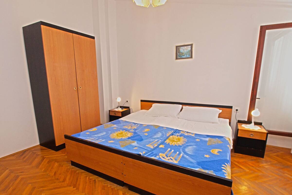 A bedroom with a wooden bed, a wardrobe, a nightstand with a lamp, and a small framed picture on the wall.