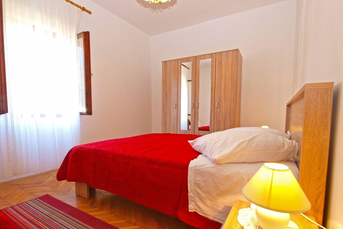 A bedroom with a red bedspread, a white pillow, a wooden wardrobe, a bedside lamp, and a striped rug.
