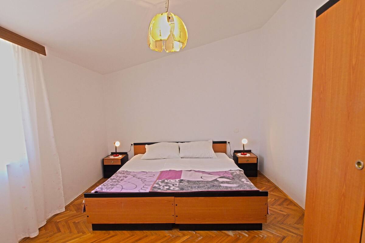 A bedroom with a wooden bed, two bedside tables with lamps, a hanging light fixture, and a wooden wardrobe.