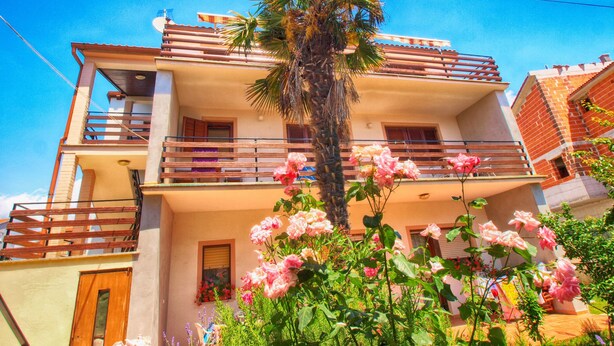 Un edificio de dos pisos con balcones, una palmera y flores rosadas.