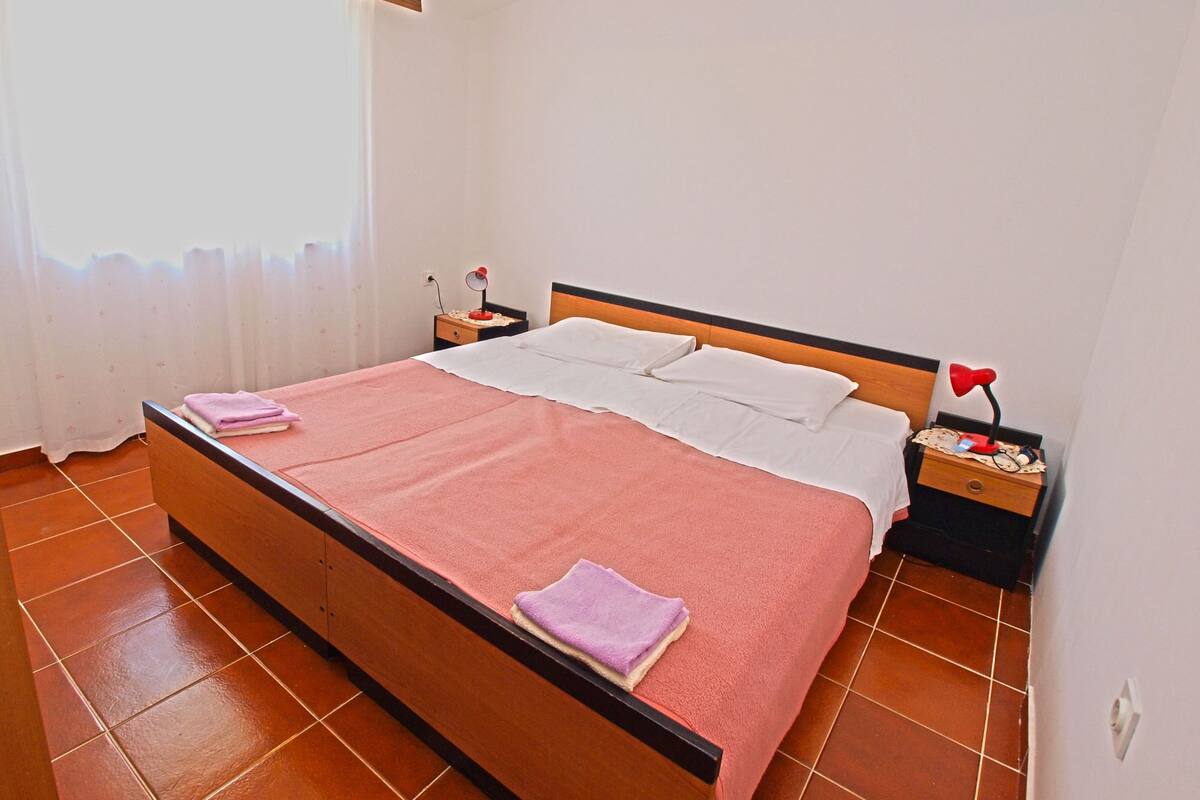 A neatly made bed with a pink blanket and white pillows, a bedside table with a lamp, and a window with curtains.