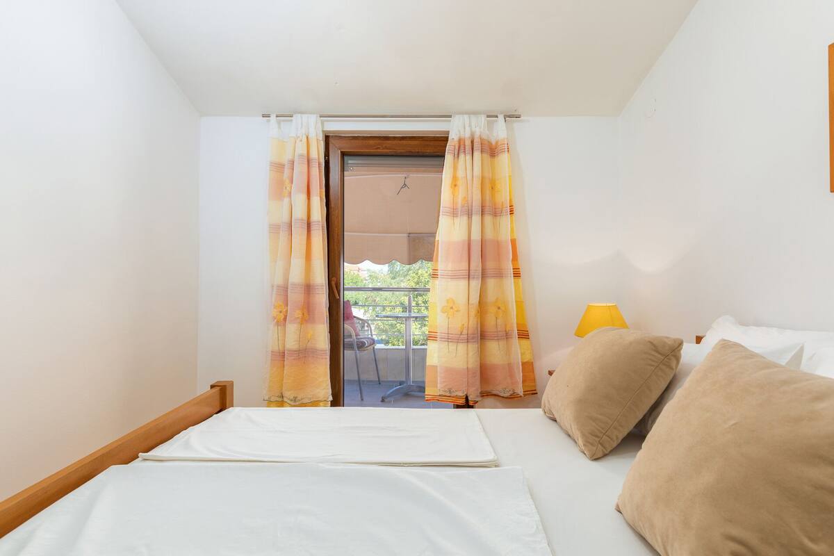 A bedroom with a bed, two pillows, a nightstand with a lamp, and a balcony visible through curtains.