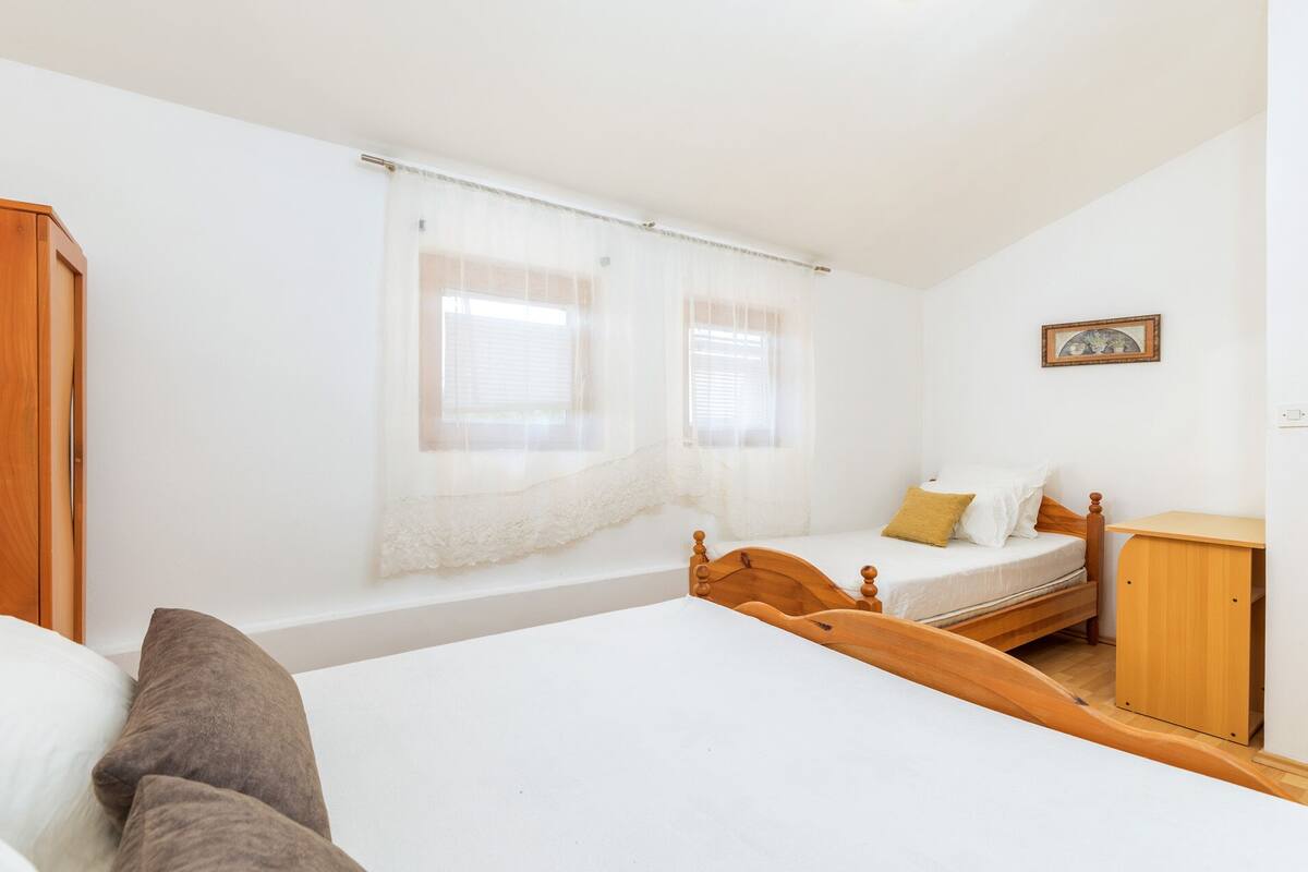 A bedroom with a large bed, a smaller bed, a wooden wardrobe, and a window with sheer curtains.