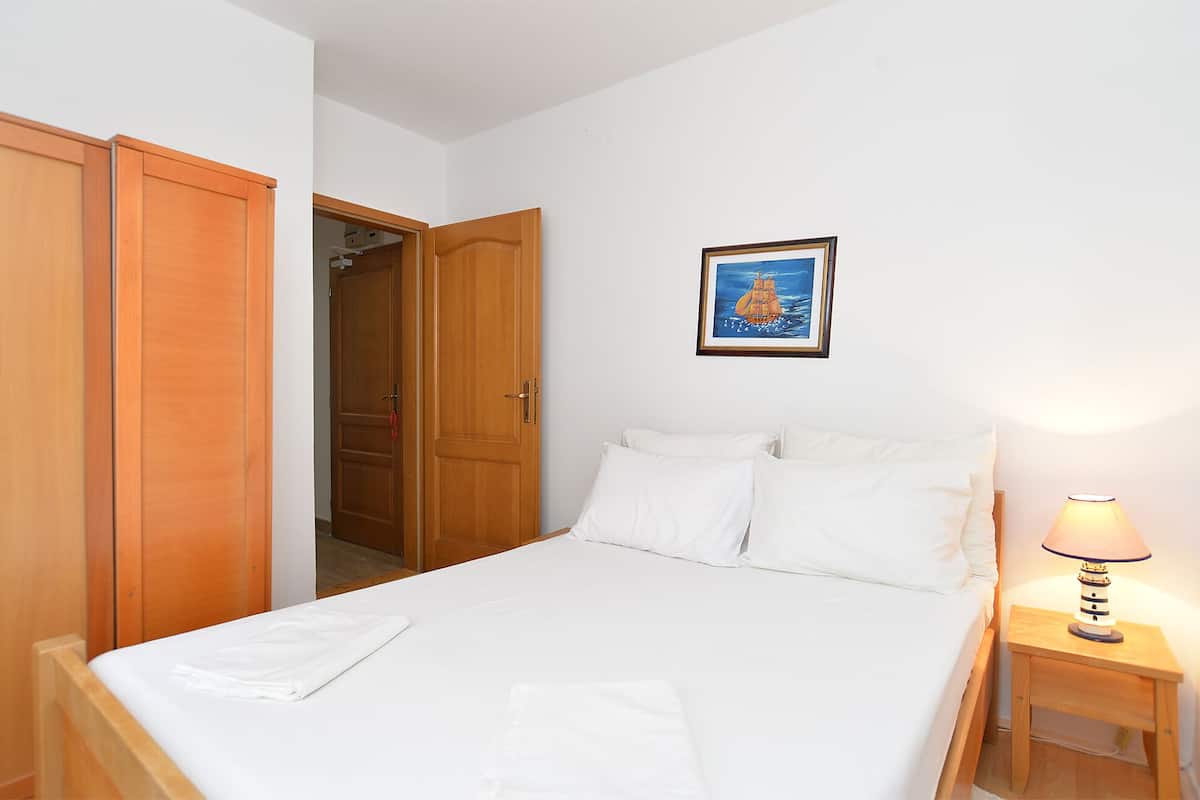 A bedroom with a bed, white linens, a wooden wardrobe, a door, a framed picture, and a bedside lamp.