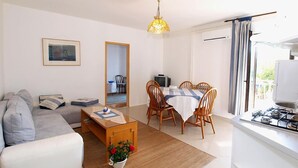 Un salón con sofá, mesa de centro y mesa de comedor con sillas. Hay una pequeña zona de cocina con estufa y una ventana con cortinas.