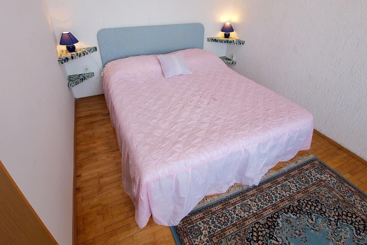 A bedroom with a single bed, two bedside tables with lamps, a patterned rug, and wooden flooring.