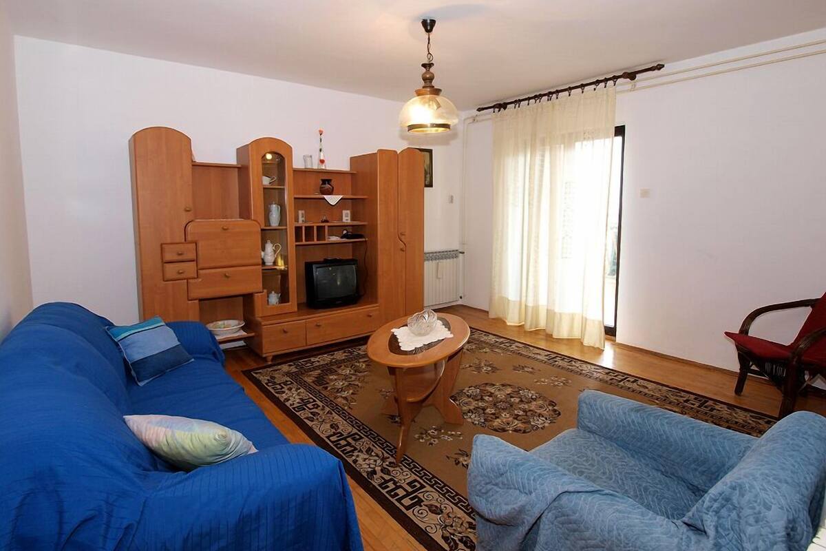 A living room with a blue sofa, a blue armchair, a wooden entertainment unit with a television, and a patterned rug.