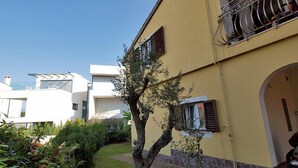 Un edificio moderno con fachada amarilla, un balcón con barandilla de metal y un pequeño árbol en primer plano.