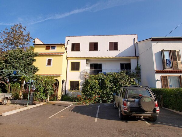 Un edificio residencial de dos pisos con fachada blanca y contraventanas marrones, un área de estacionamiento y un coche aparcado.