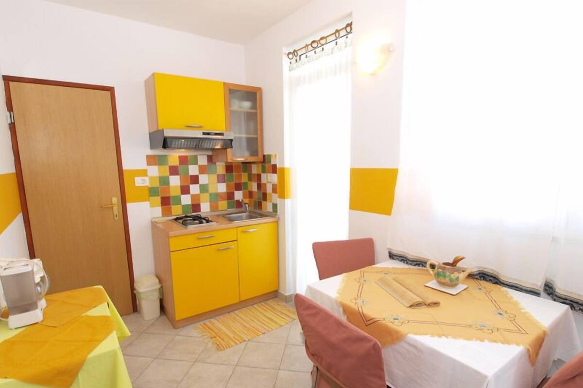 A compact kitchen with yellow cabinets, a gas stove, and a sink. There's a dining table with a yellow tablecloth and chairs, and a window with curtains.