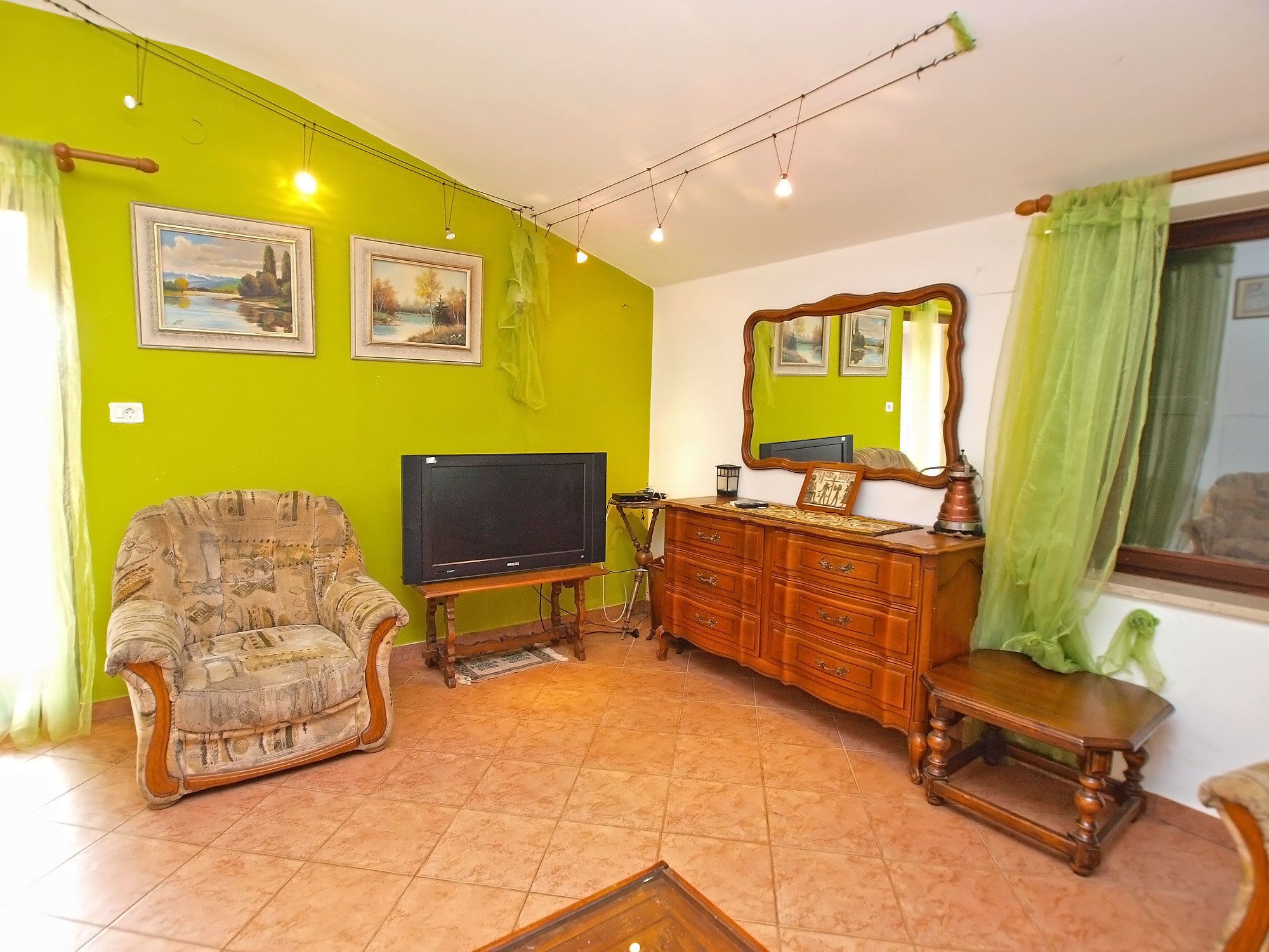 A living room with a green wall, a television, a wooden dresser, a chair, and a mirror.