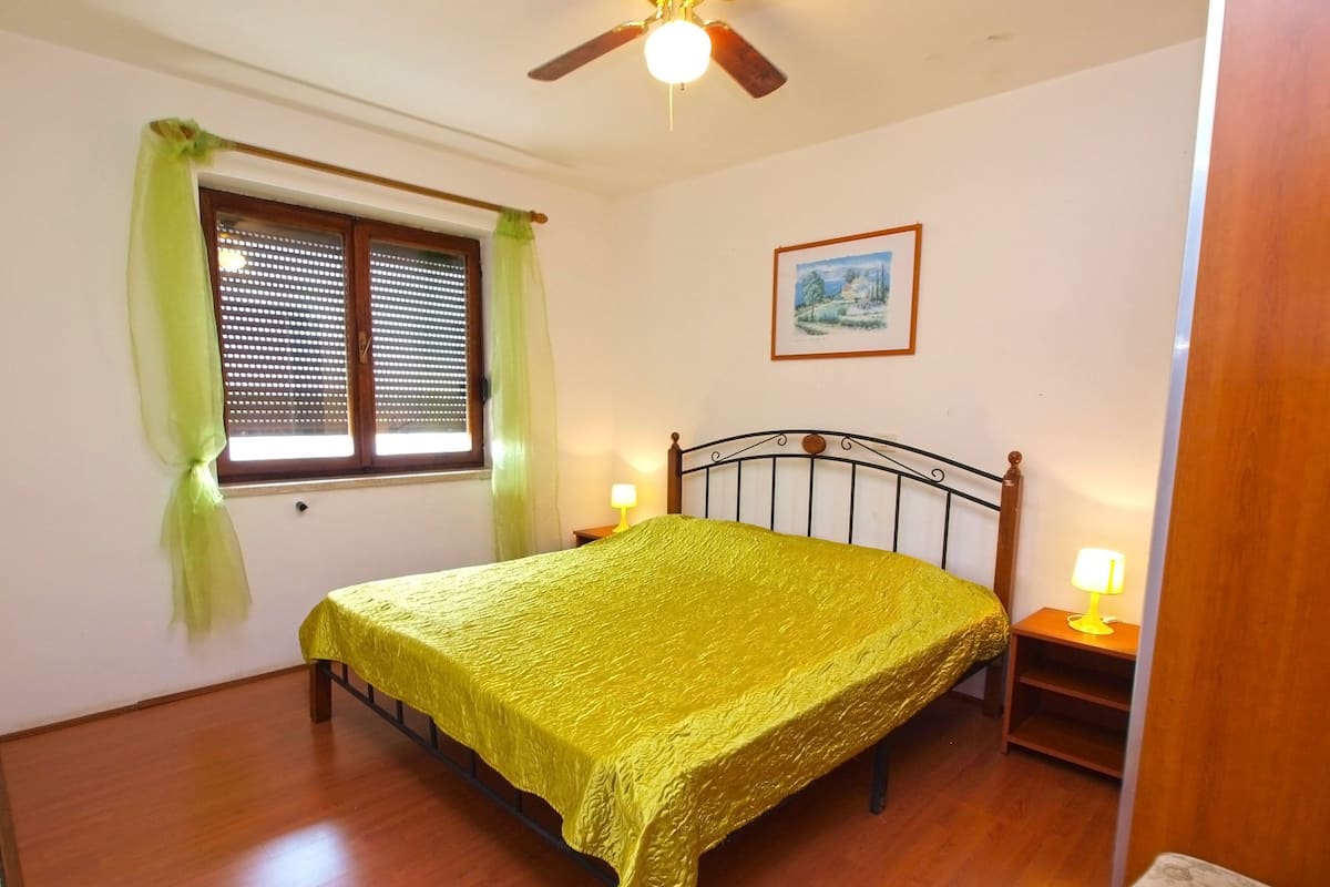 Une chambre à coucher comprenant un lit, deux lampes de chevet, un ventilateur de plafond, une fenêtre avec des stores et un tableau encadré accroché au mur.