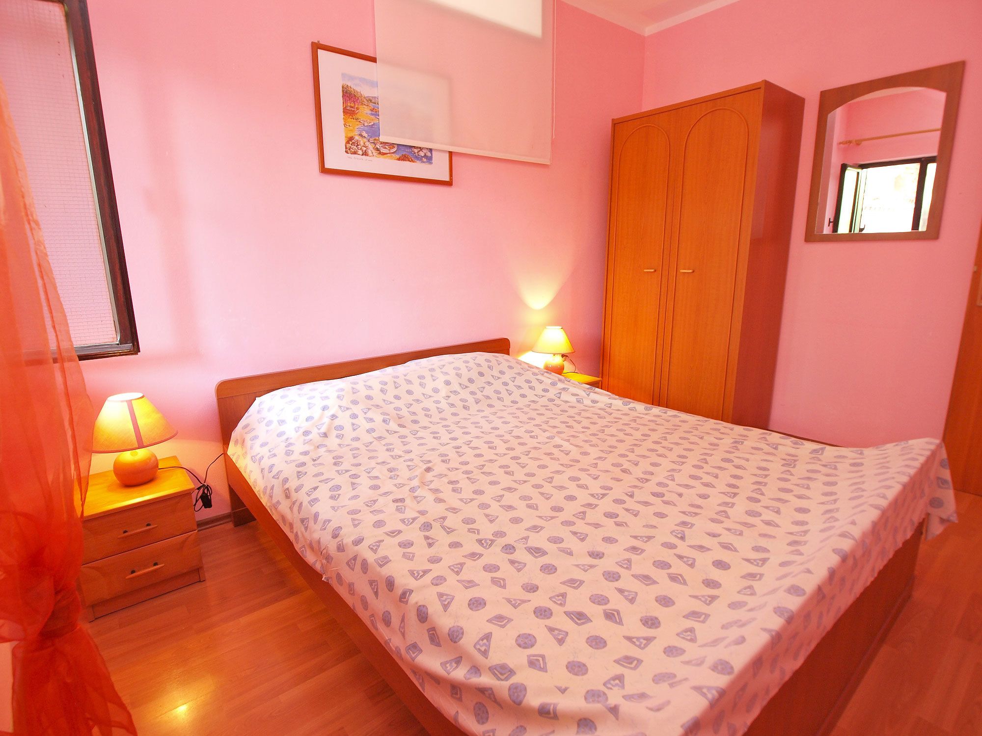 A bedroom with a bed, a wooden wardrobe, a bedside table with a lamp, and a window with curtains.