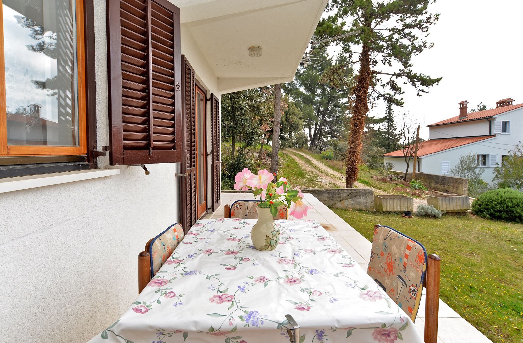 A table set outdoors with a floral tablecloth, a vase with flowers, and wooden chairs.
