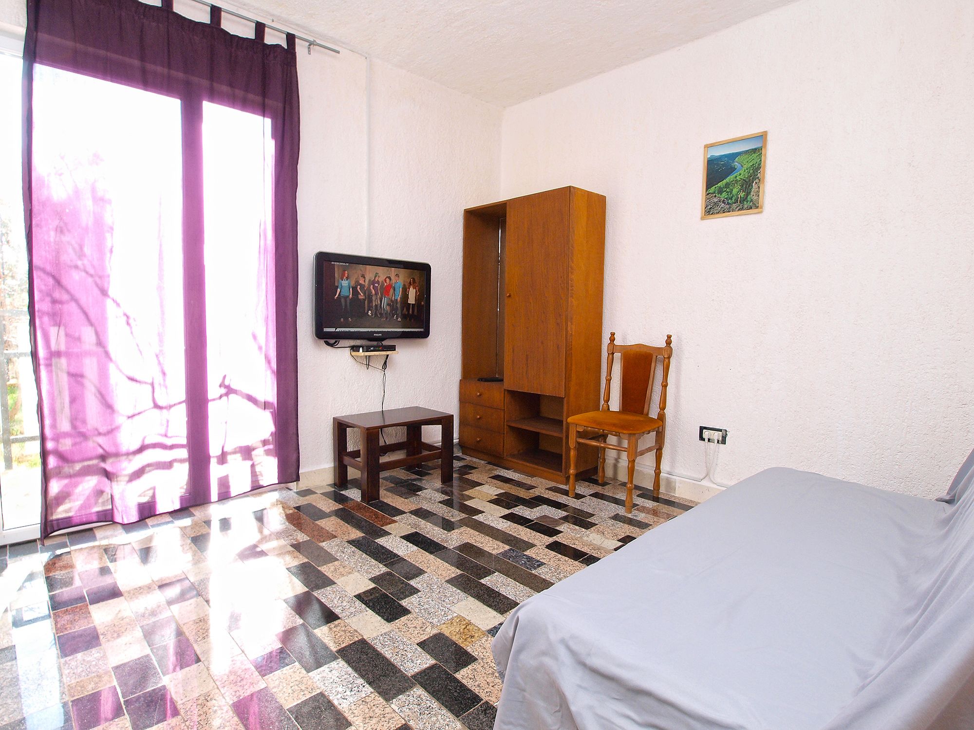 Ein Hotelzimmer mit Schachbrettmuster-Boden, einem Bett, einem Fernseher, einem Holzschrank und einem Sessel.