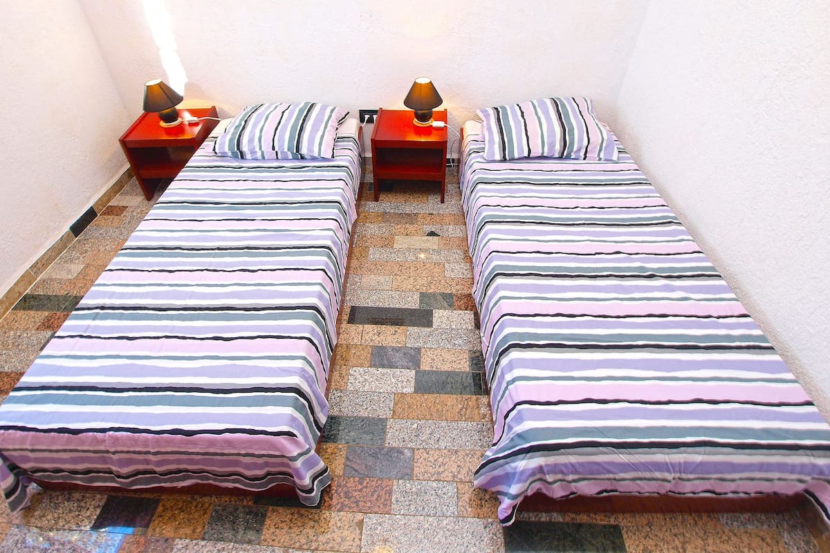 A corridor with four single beds aligned against the wall, each with a striped bedspread and a small bedside table with a lamp.