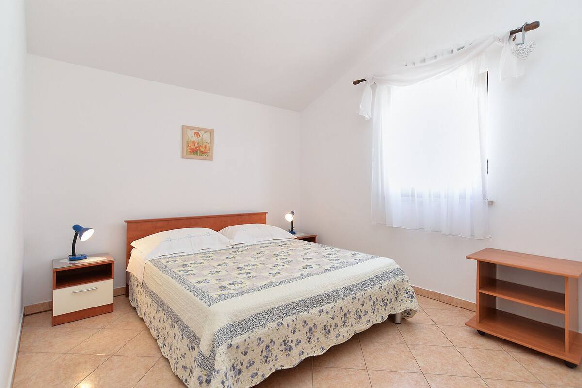 A bedroom with a bed, two bedside tables, a nightstand, a window with sheer curtains, and a small wooden shelf.
