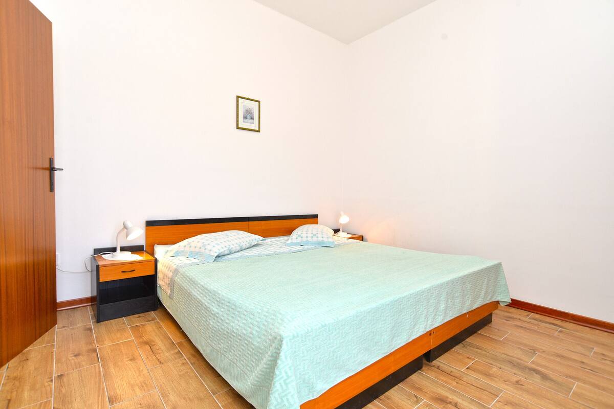 A bedroom with a bed, two bedside tables, a nightstand, a wooden door, and a framed picture on the wall.