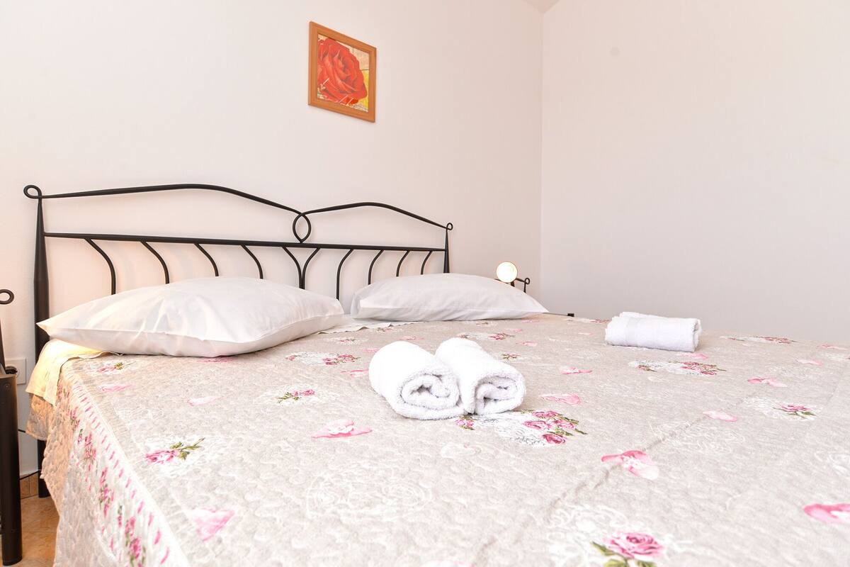 A neatly made bed with a floral bedspread, two rolled towels, and a single pillow.
