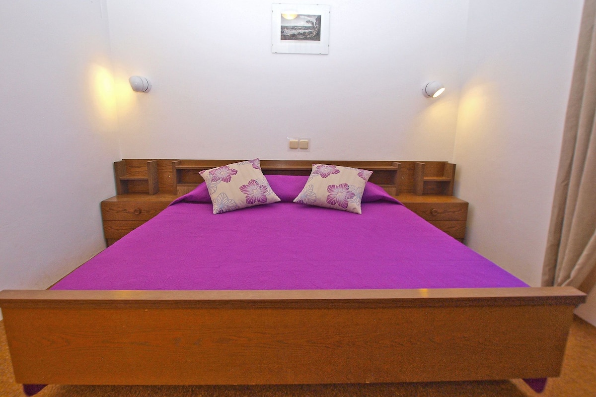 Habitación individual con una cama de madera, ropa de cama morada, dos almohadas florales, mesitas de noche e iluminación empotrada en la pared.