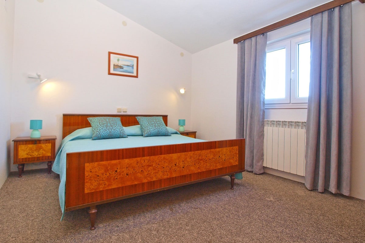 Un dormitorio con cama de madera, dos mesitas de noche, una mesita auxiliar, una ventana con cortinas y un cuadro colgado en la pared.