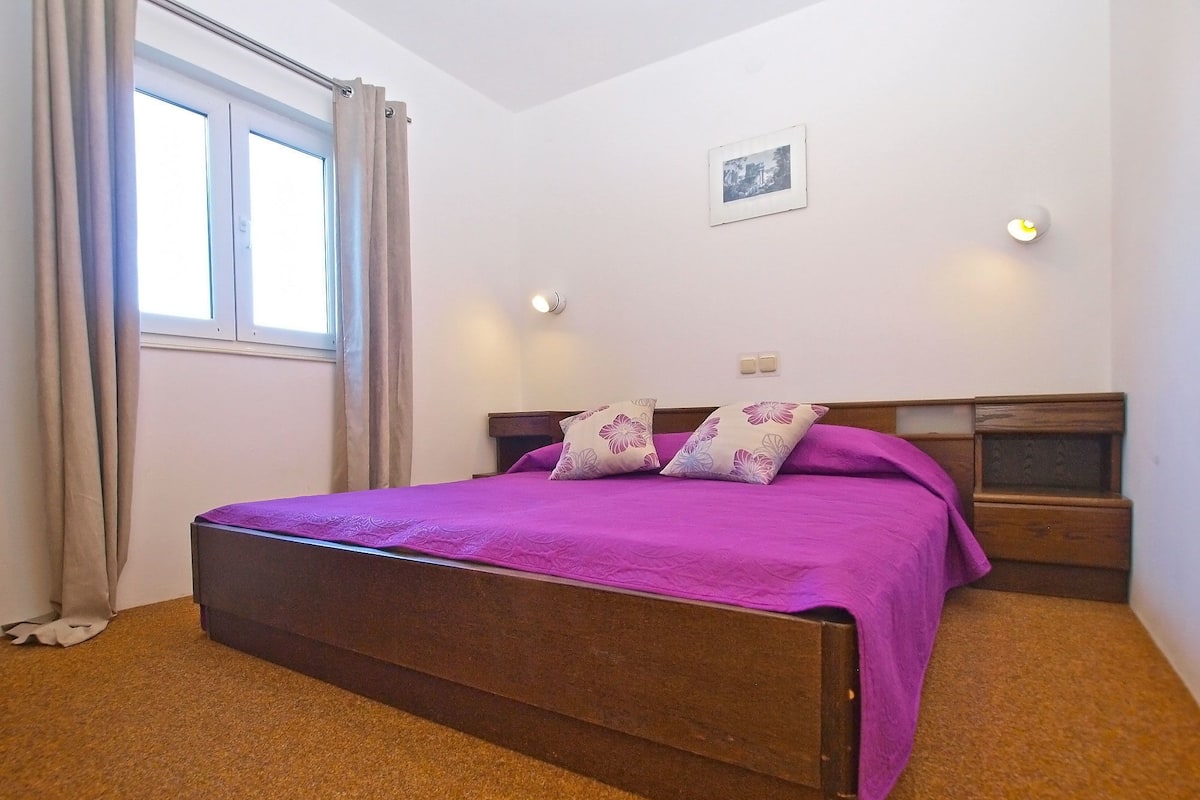 Un dormitorio con cama de madera, mesita de noche, lámpara de pared y ventana con cortinas.