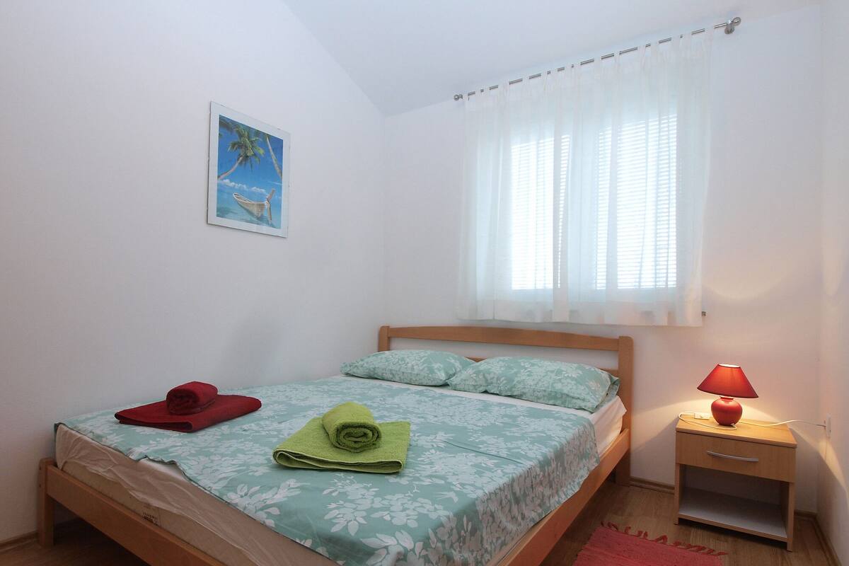 A single bed with a floral bedspread, a red towel, and a green towel on the bed. There is a nightstand with a red lamp beside the bed. A window with white curtains is visible, and a painting of a tropical scene hangs on the wall.