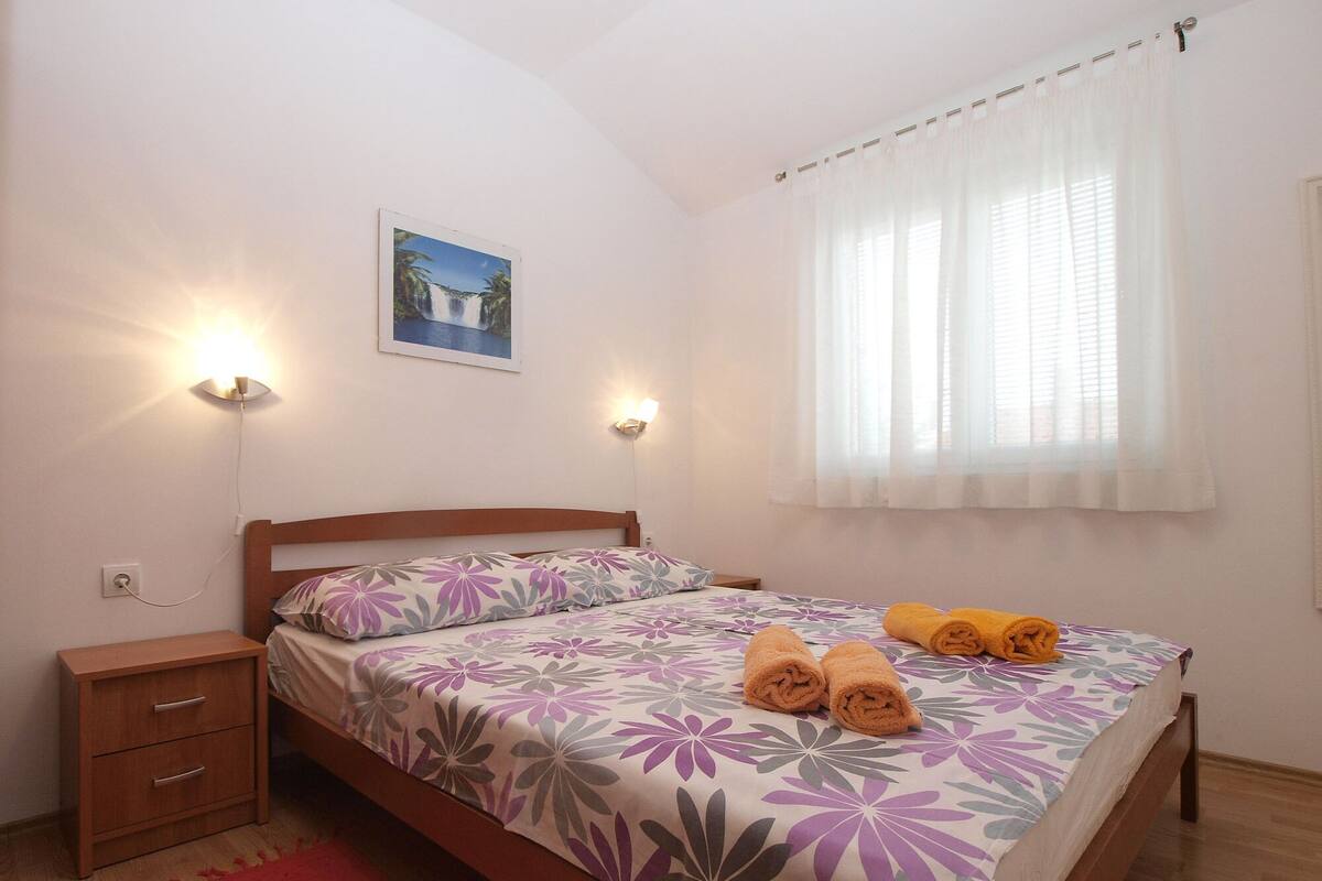 A neatly made bed with floral bedding, two bedside lamps, a nightstand, a red rug, and a window with sheer curtains.