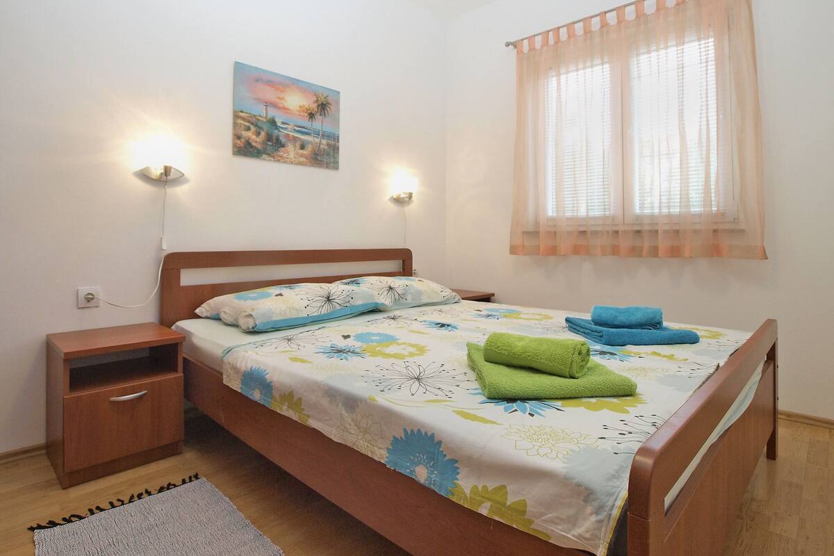 A bedroom with a wooden bed, a nightstand, a bedside table, a window with sheer curtains, and a painting on the wall.