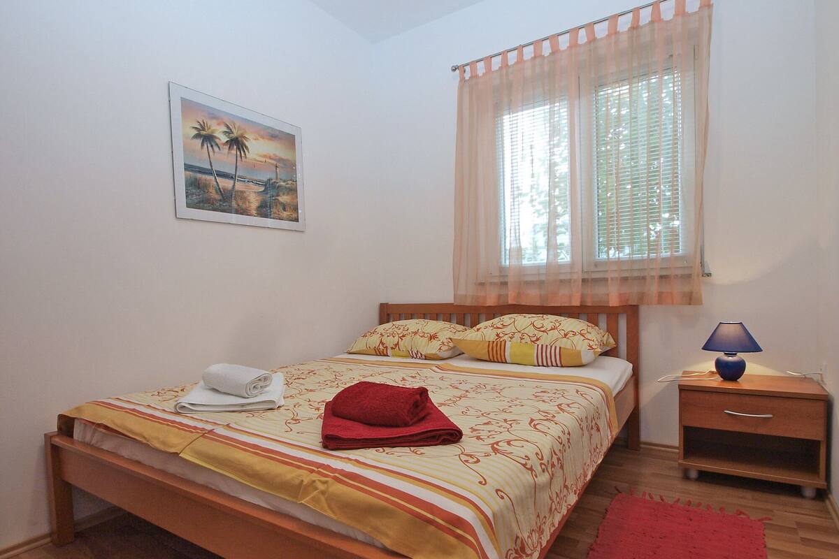 A bedroom with a bed, a nightstand, a lamp, a window with sheer curtains, and a painting on the wall.
