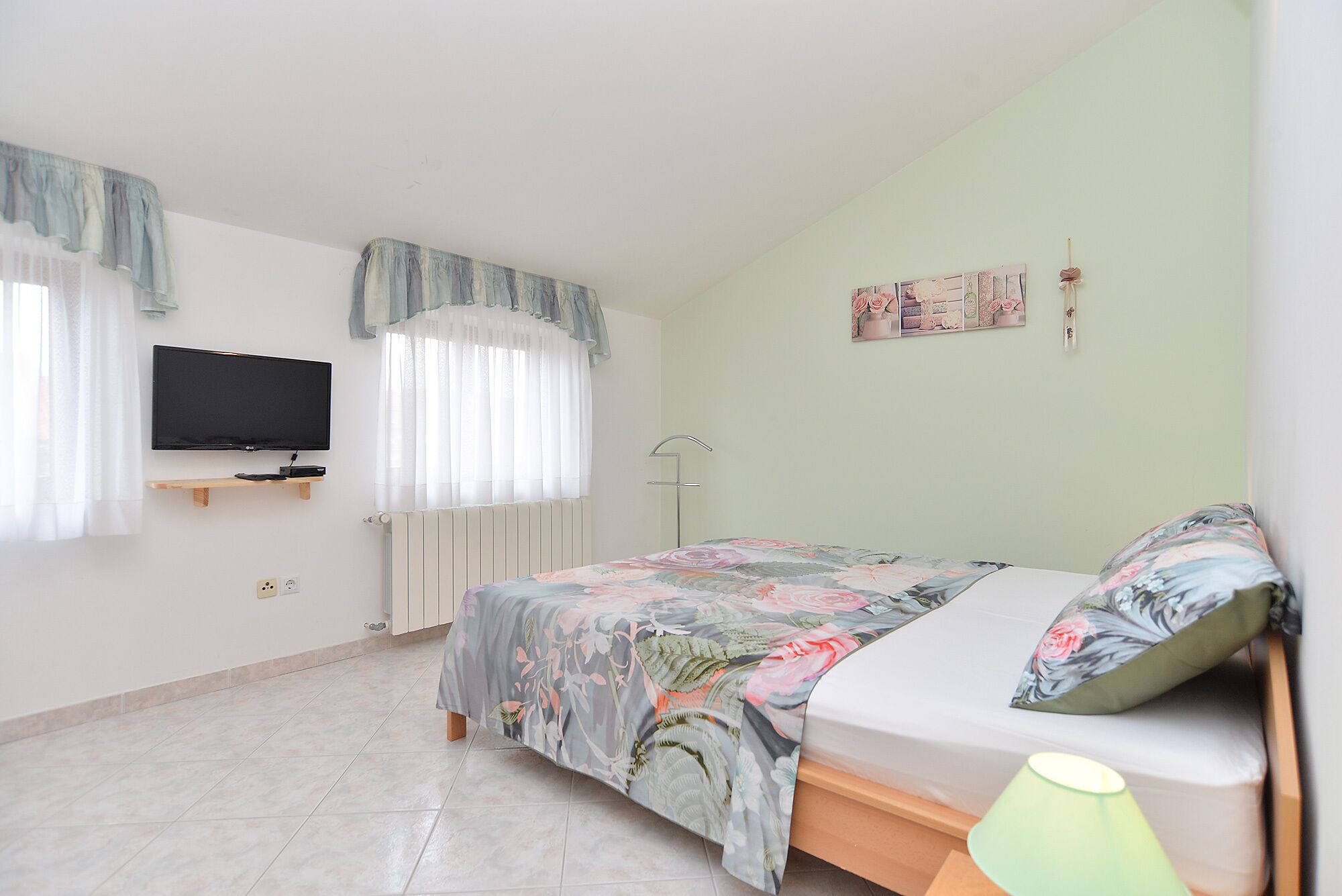 A bedroom with a bed, a TV mounted on the wall, a radiator, and a window with curtains.