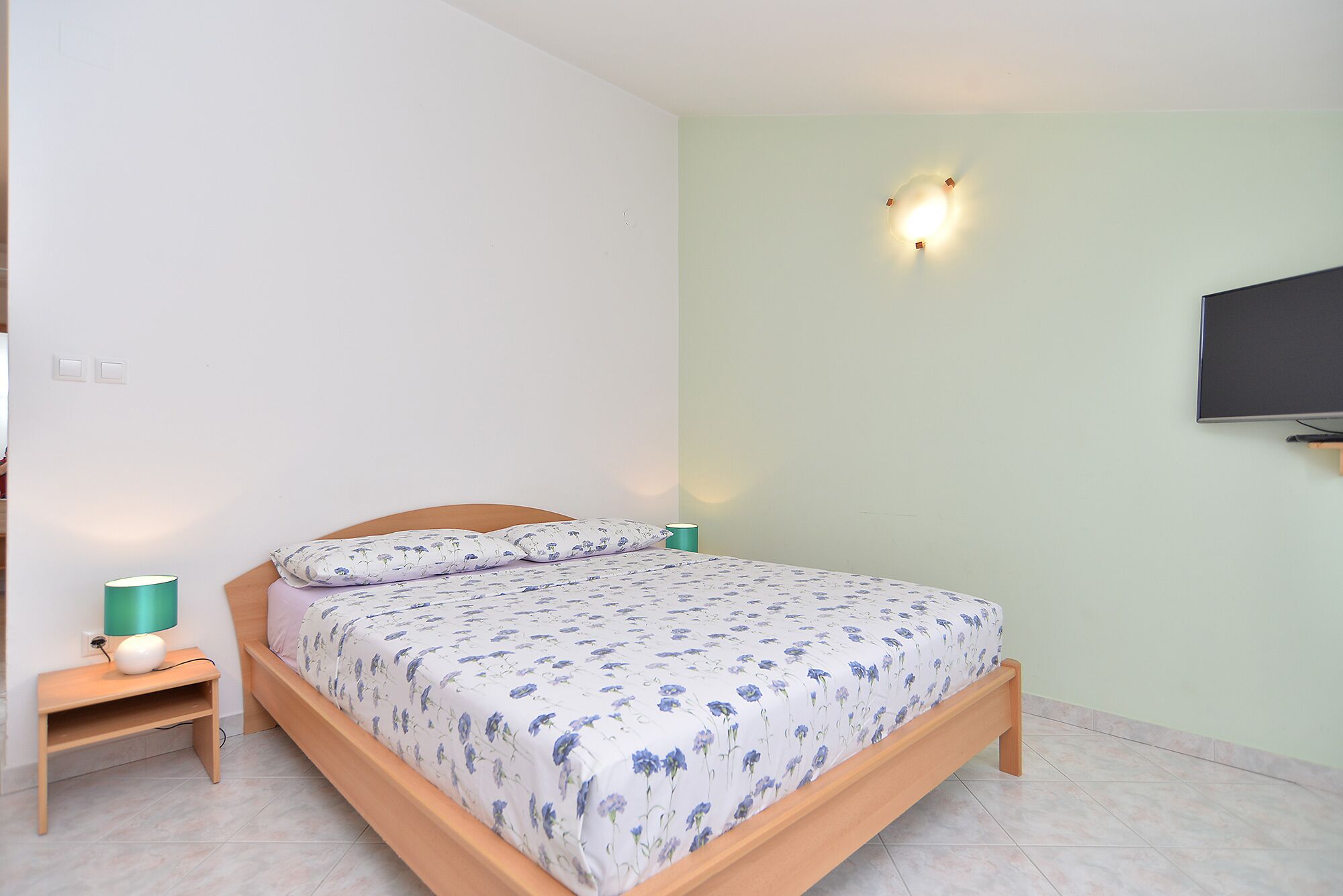 A bedroom with a bed, a nightstand, a lamp, a wall-mounted TV, and a wall light.