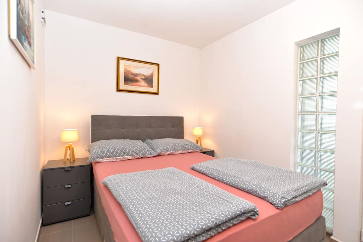 Un dormitorio con una cama, dos mesitas de noche, una lámpara de noche, una ventana con persianas y un cuadro en la pared.