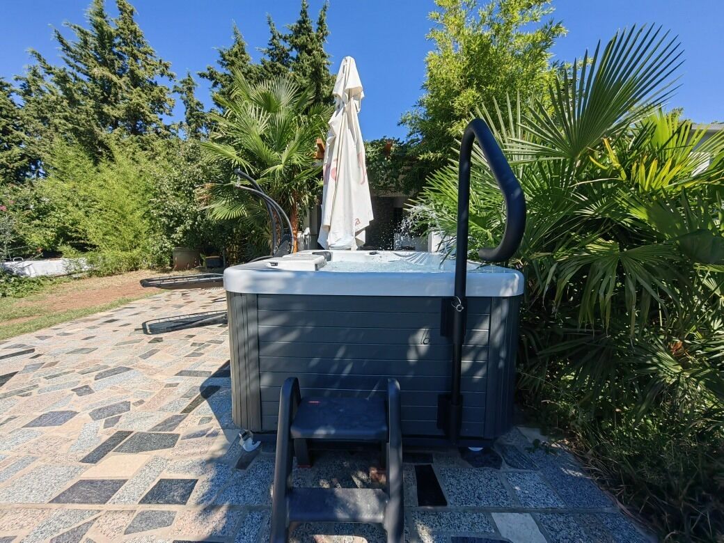 A hot tub with a ladder, surrounded by greenery and a tiled patio.