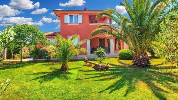 A two-story house with a red exterior, surrounded by a well-maintained garden with palm trees and a lawn. - Apartment Tatjana 411 (Medulin)