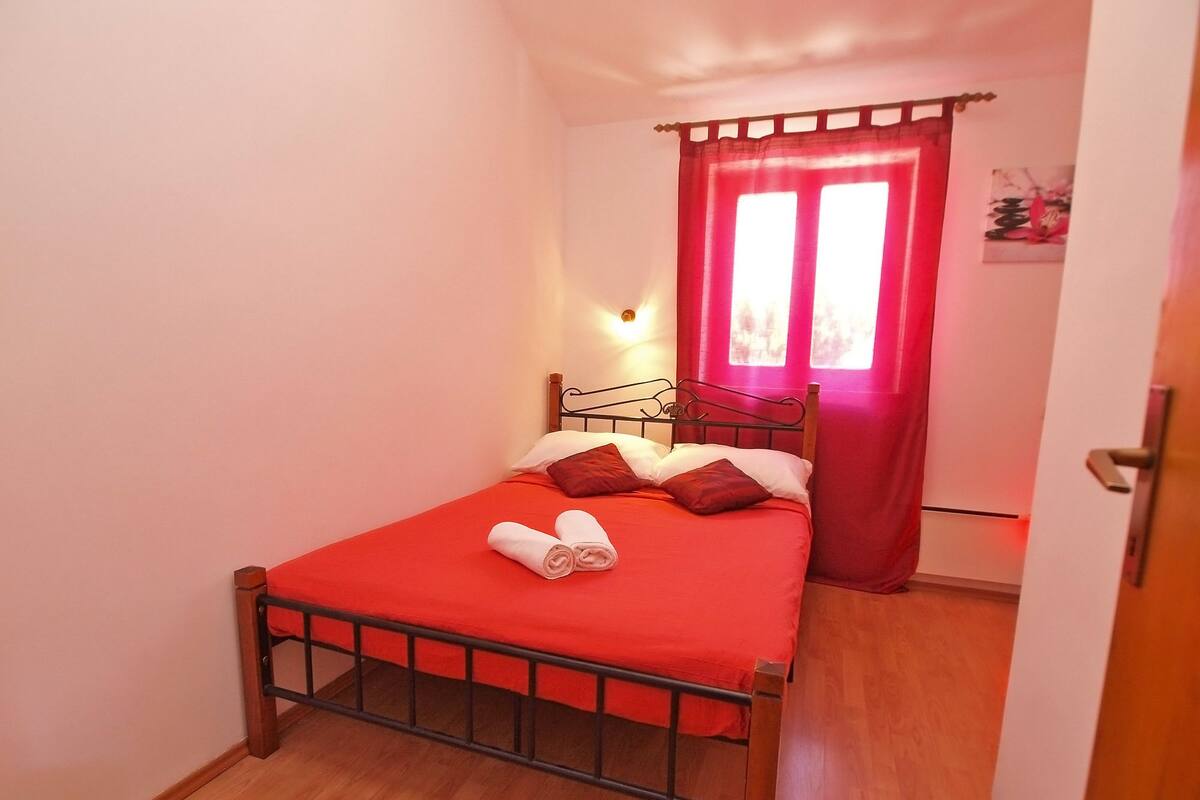 Una habitación pequeña con cama individual, cubierta roja, estructura metálica y tablero de cabecera de madera, además de una cortina roja.