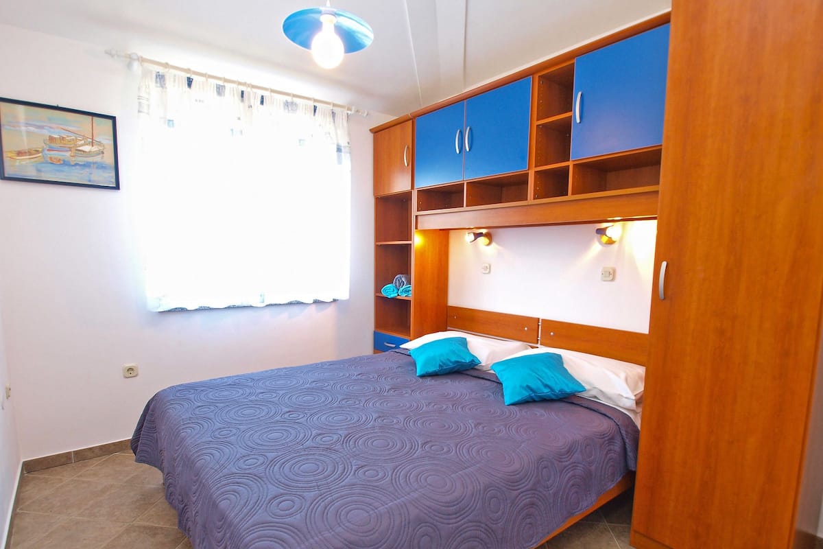 Un dormitorio con cama, almohadones azules, un armario de madera y una ventana con cortinas.