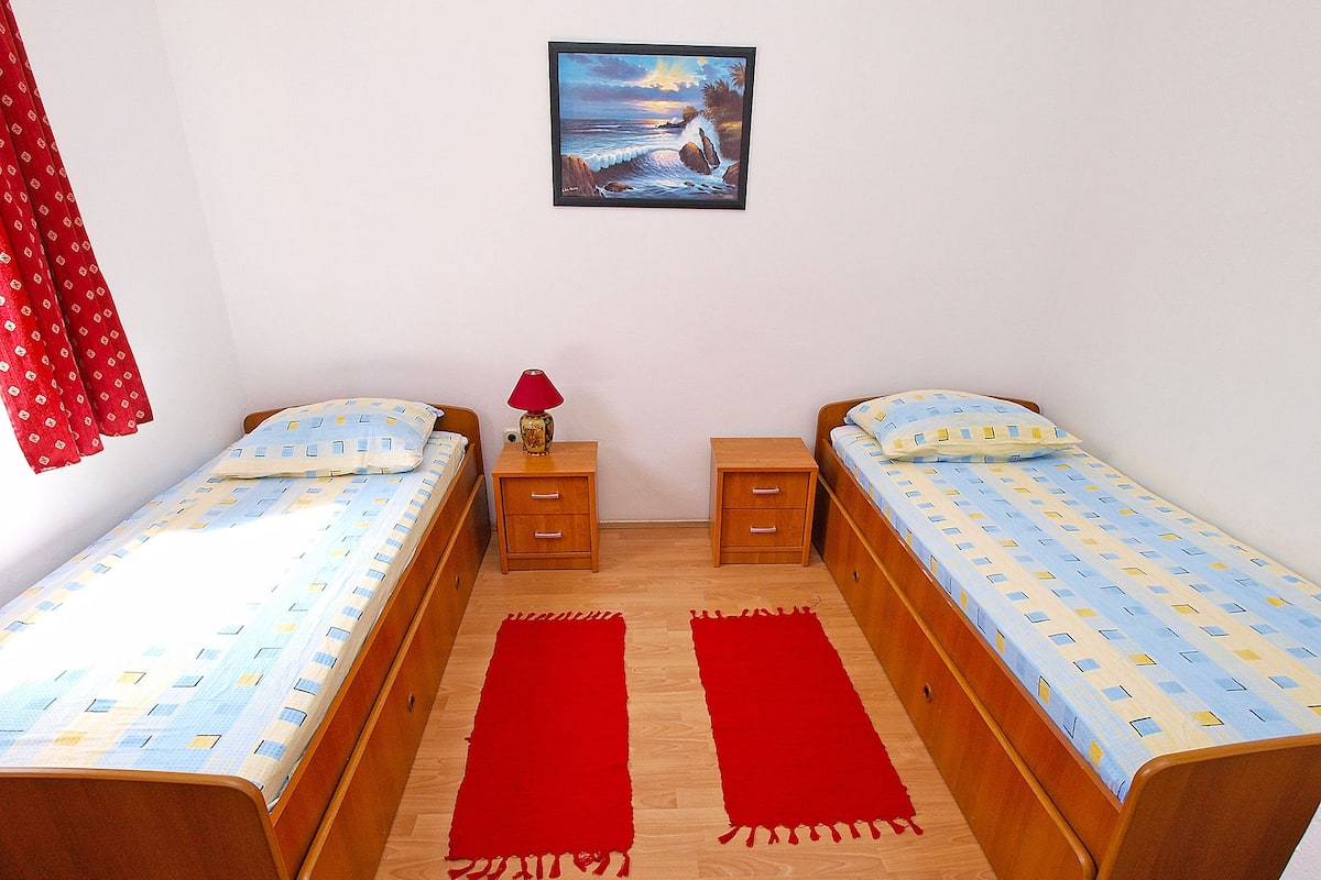 Een kamer met twee eenpersoonsbedden, een nachtkastje met een lamp, een rood vloerkleed en een schilderij aan de muur.